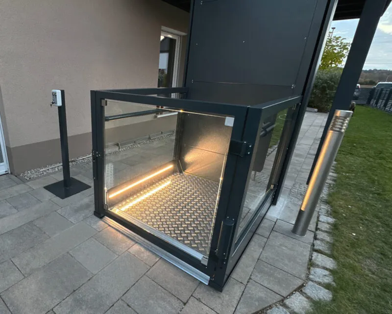 Mørk løfteplattform med glassvegger, dorkplategulv og LED-lys på terrasse utenfor bolig